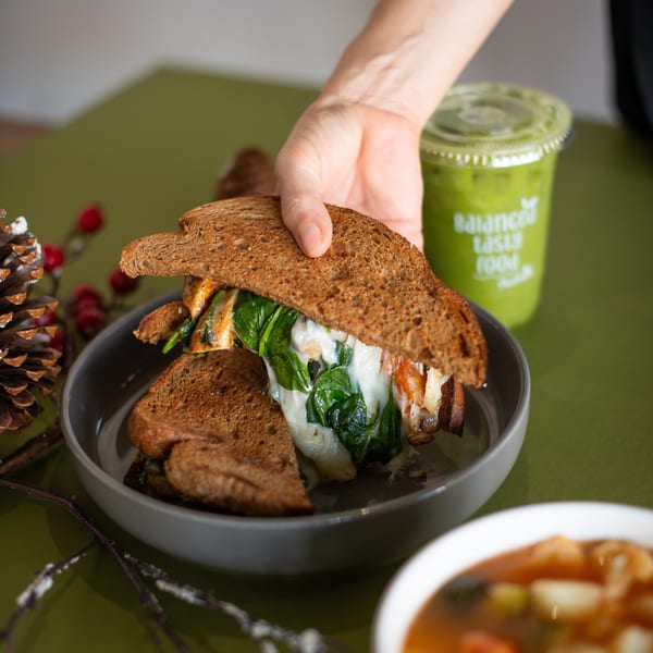 Hand reaching for a toasted sandwich with melted cheese and spinach, next to a green smoothie.