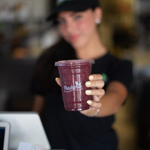 A Fresheria employee in a black cap and shirt holds out a berry smoothie.