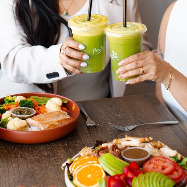 Two women toasting green Fresheria smoothies over plates of salmon and salad.