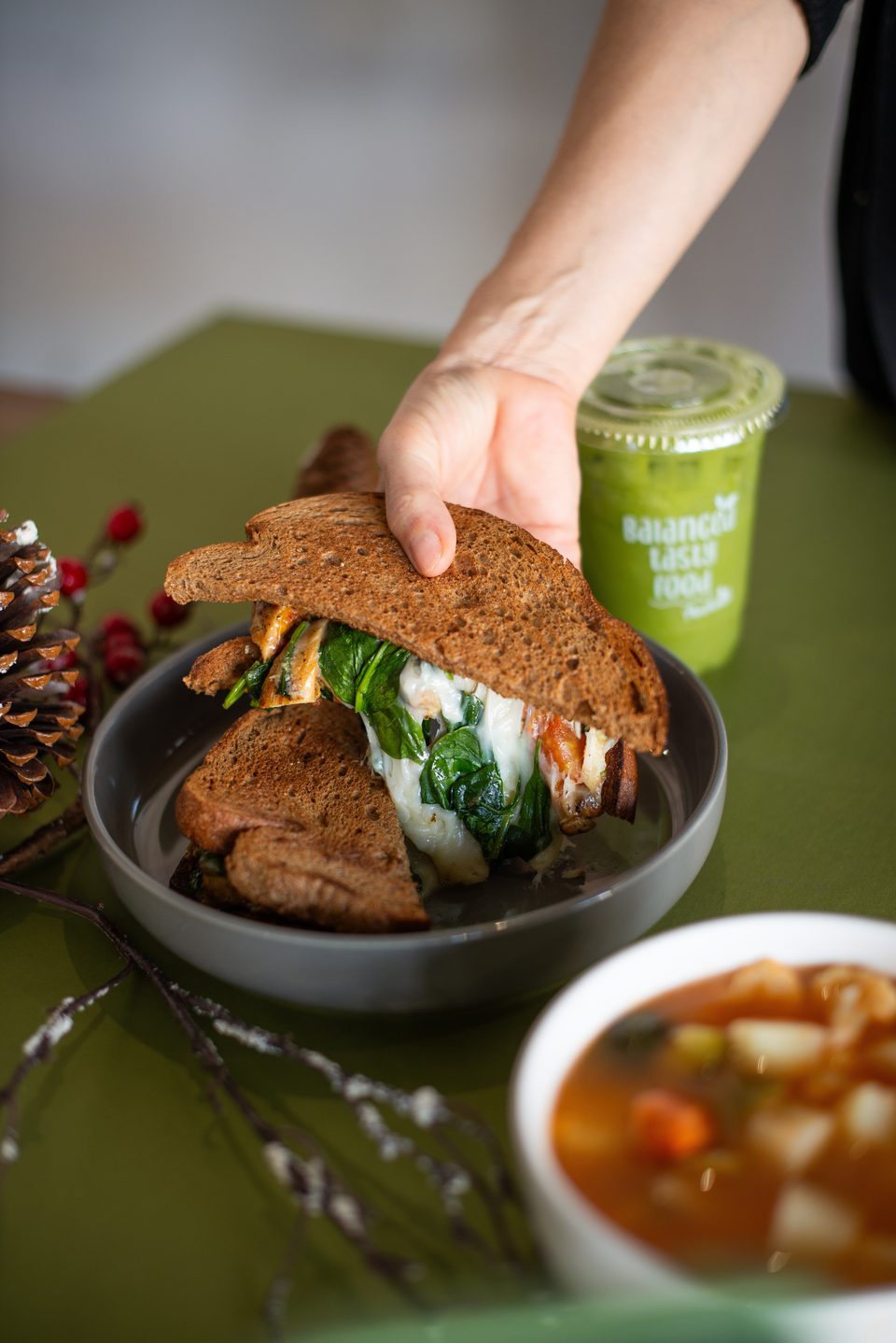 A hand reaching for a grilled cheese sandwich with spinach, next to a green smoothie and soup.