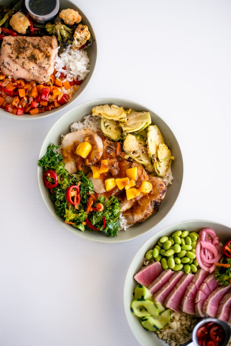 Three colorful bowls of food, including salmon, pork with mango, and tuna with edamame.