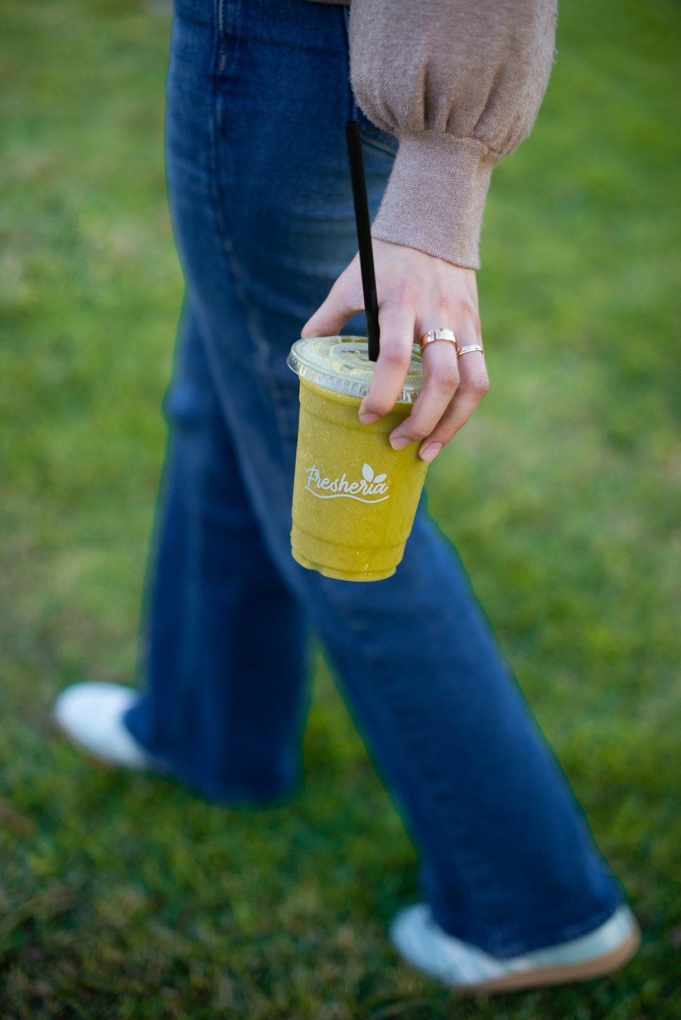 Person in jeans and a sweater holding a green Fresheria smoothie with a black straw.