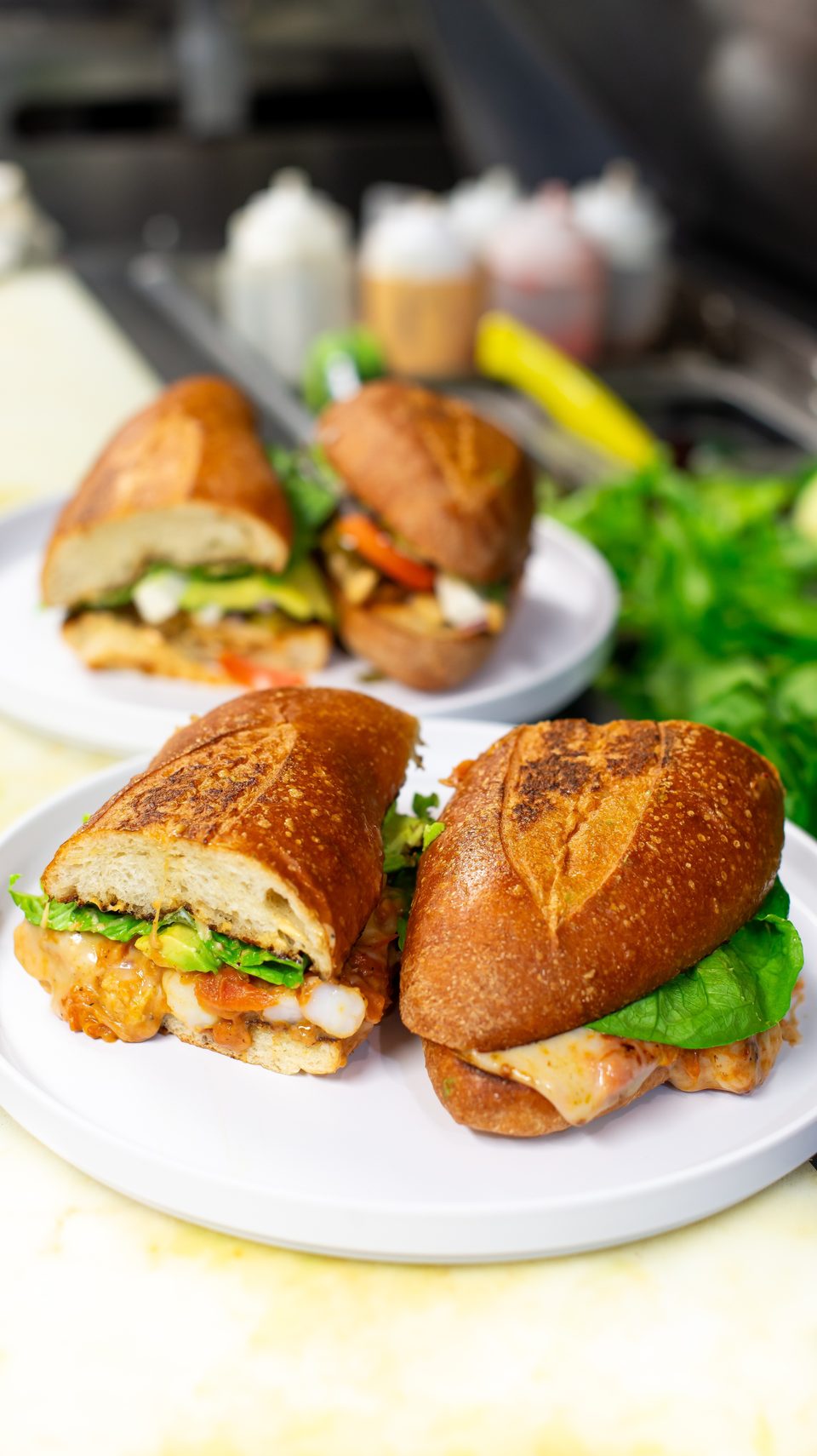Two shrimp po'boy sandwiches with lettuce, avocado, and sauce on white plates in a kitchen.