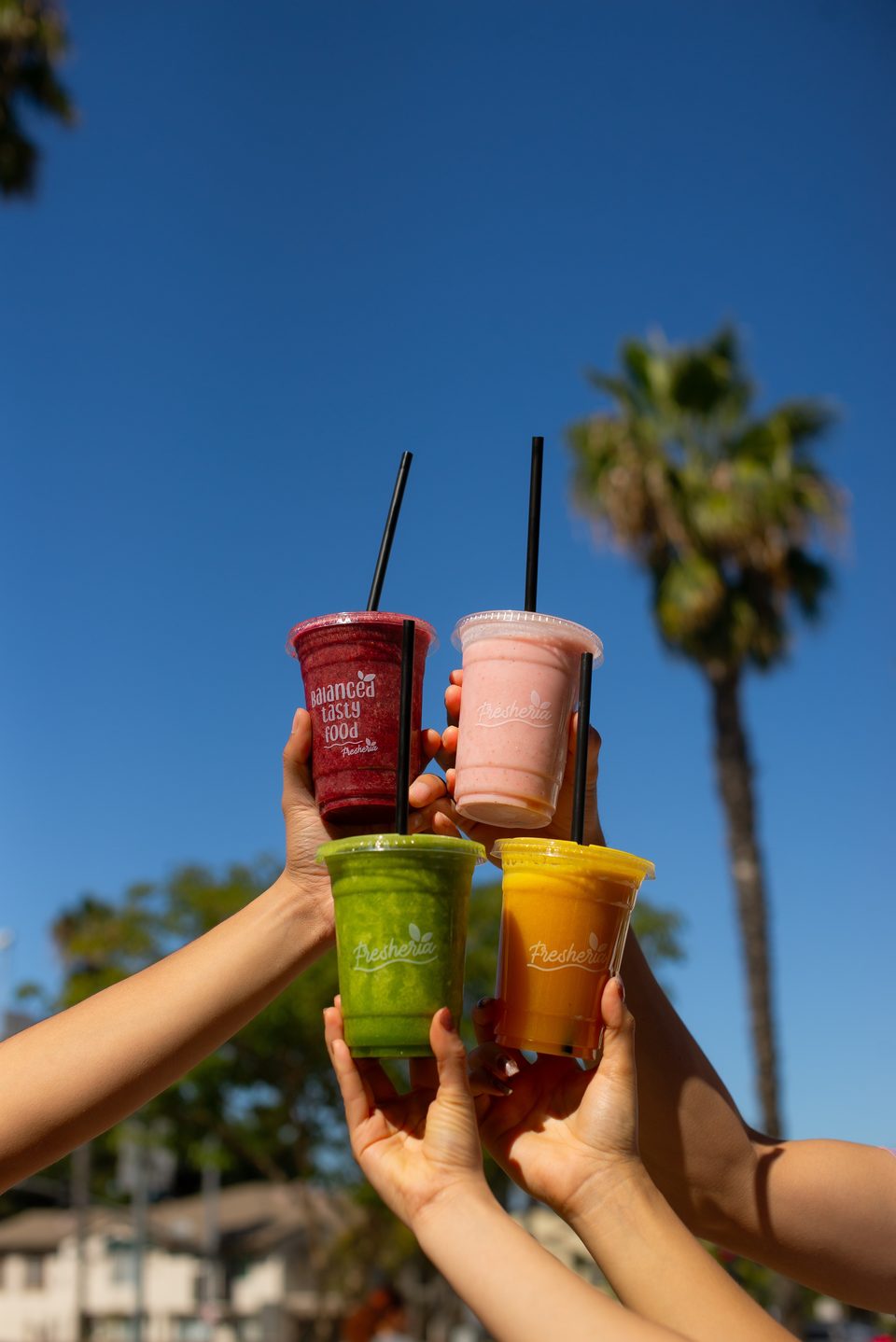 Four hands hold up colorful Fresheria smoothies against a clear blue sky with palm trees.