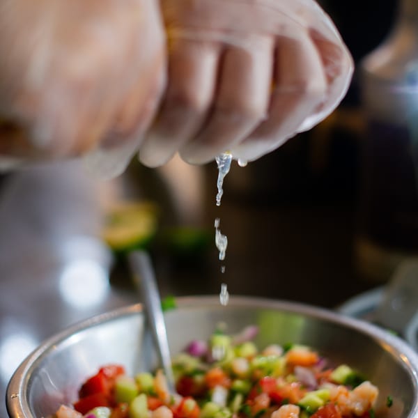 Gloved hands squeeze a lime over a bowl of fresh shrimp ceviche with diced vegetables.