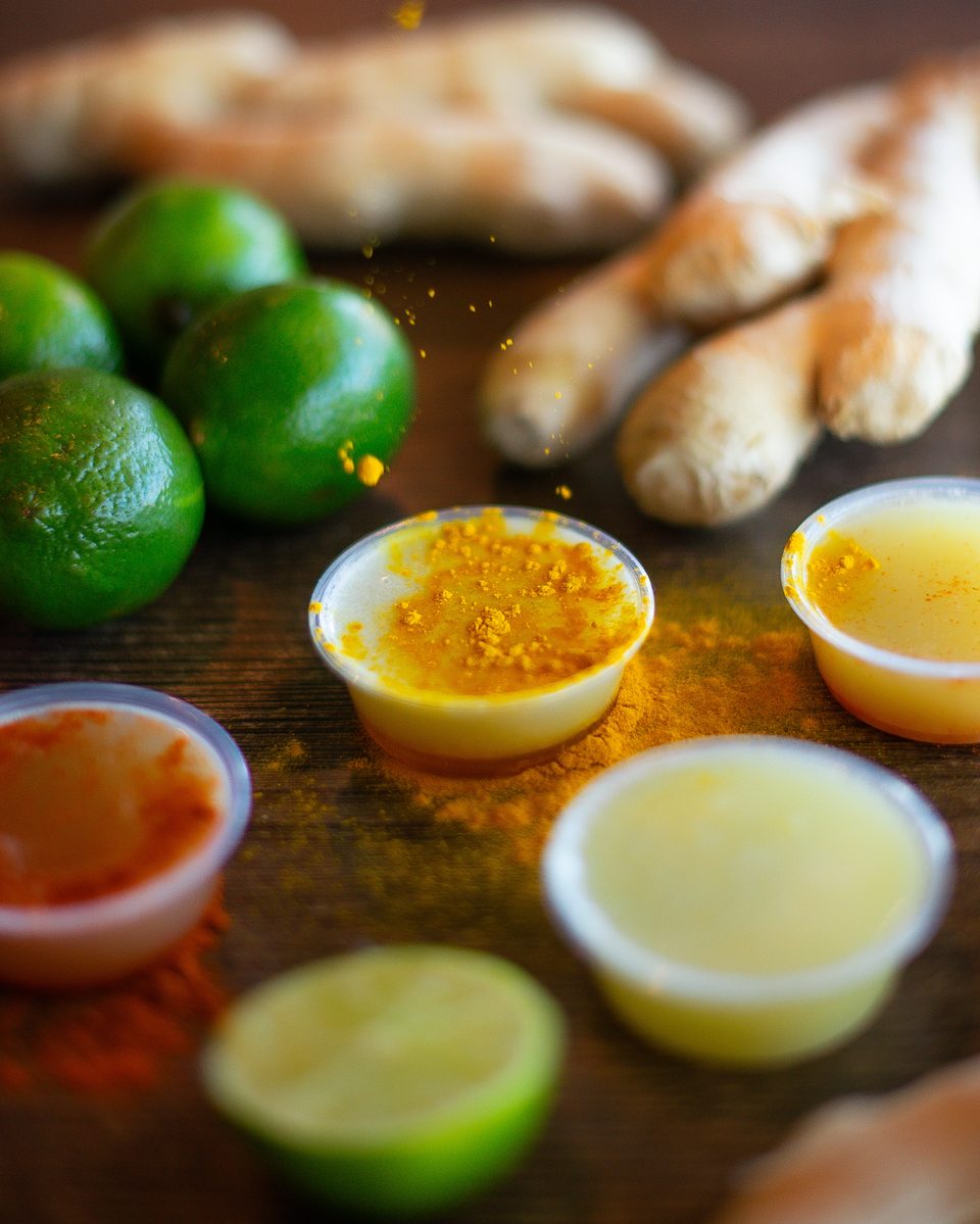 Limes, ginger, and small cups of yellow and orange spices and liquids on a wooden surface.