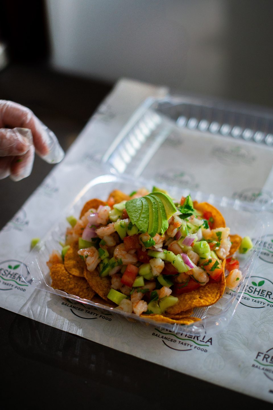 A chef's gloved hand reaches towards a plastic container of shrimp ceviche with avocado and tortilla chips.