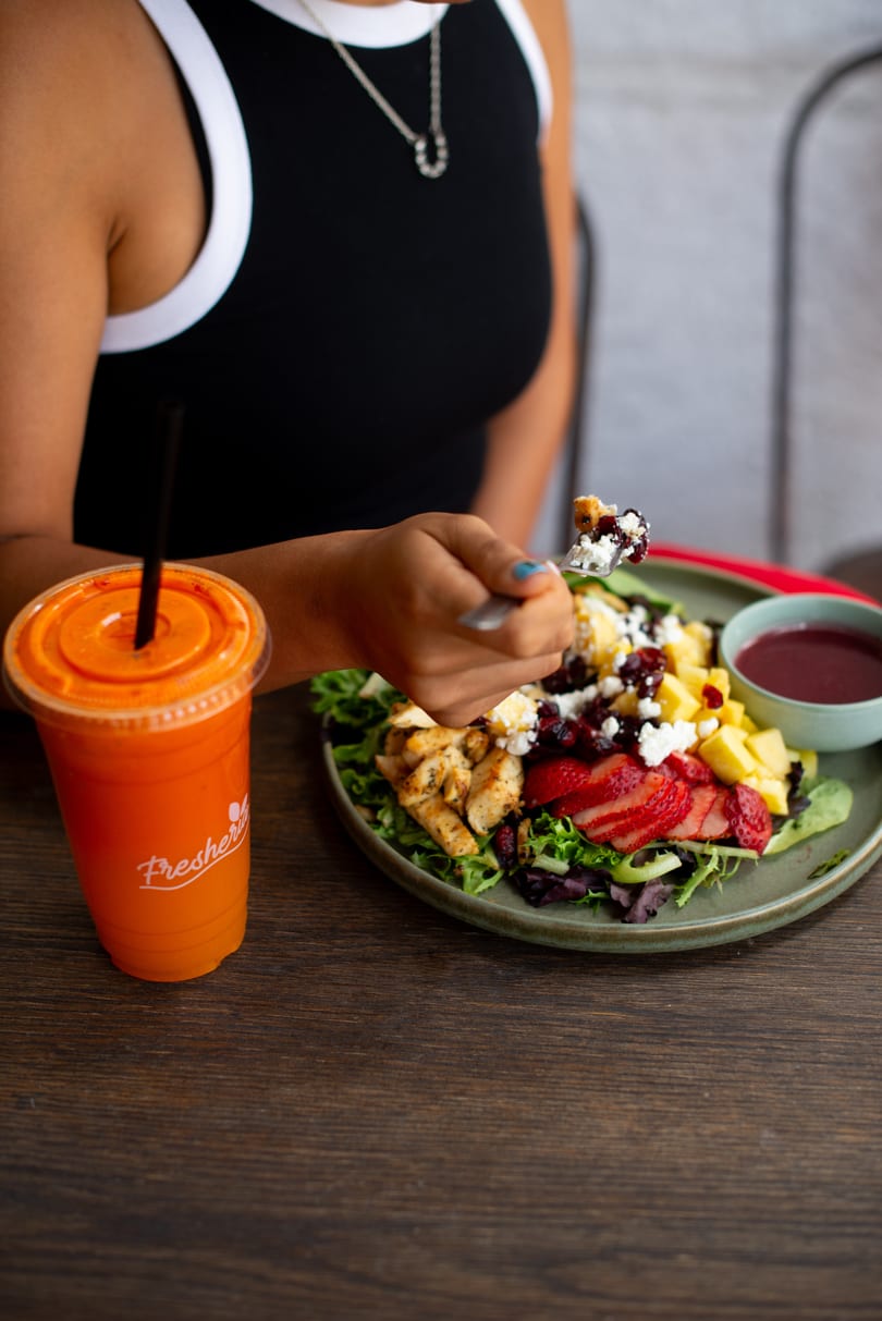 A person eating a colorful salad with chicken, fruit, and feta, next to a Fresheria juice.