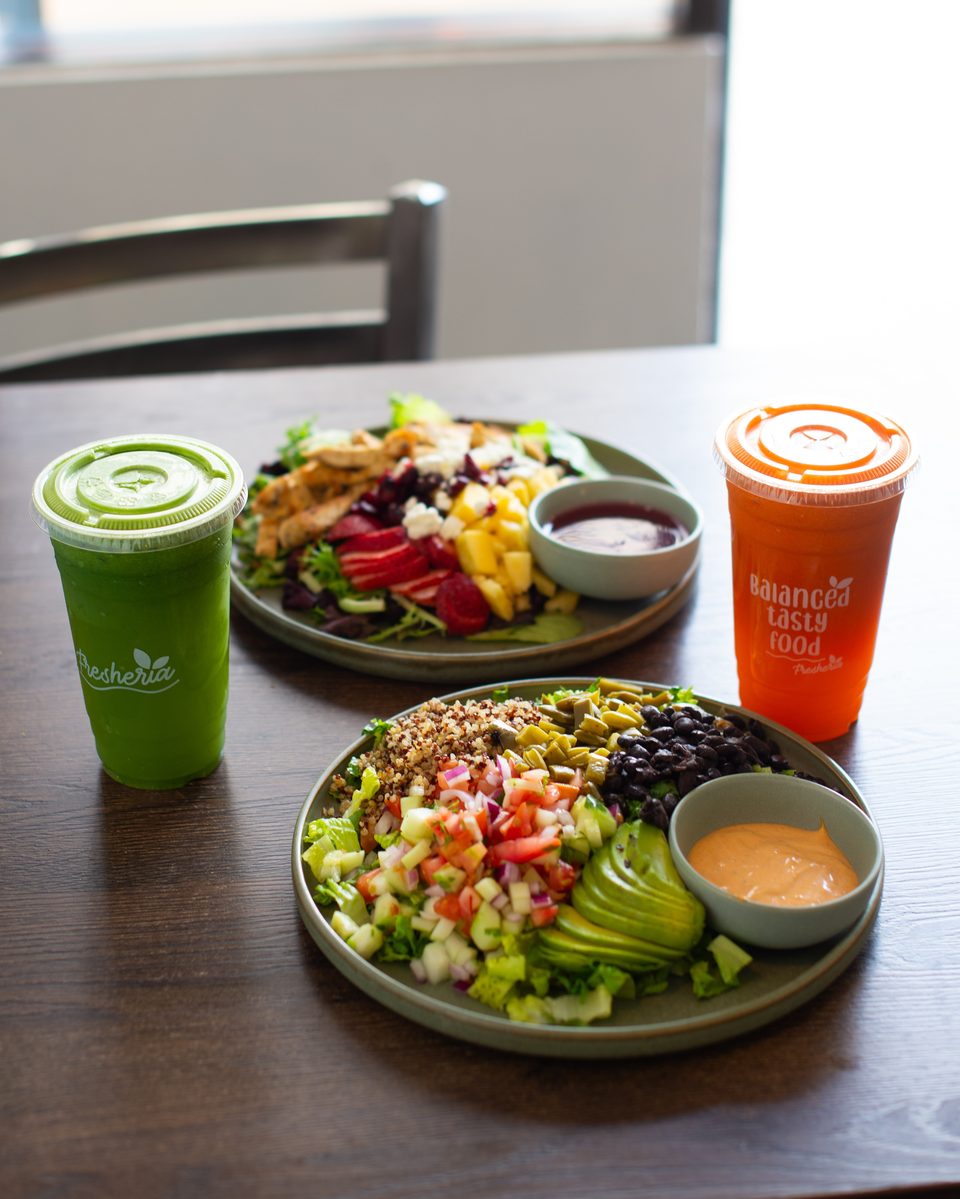 Two vibrant salads and two Freshteria drinks, a green juice and an orange juice, on a wooden table.