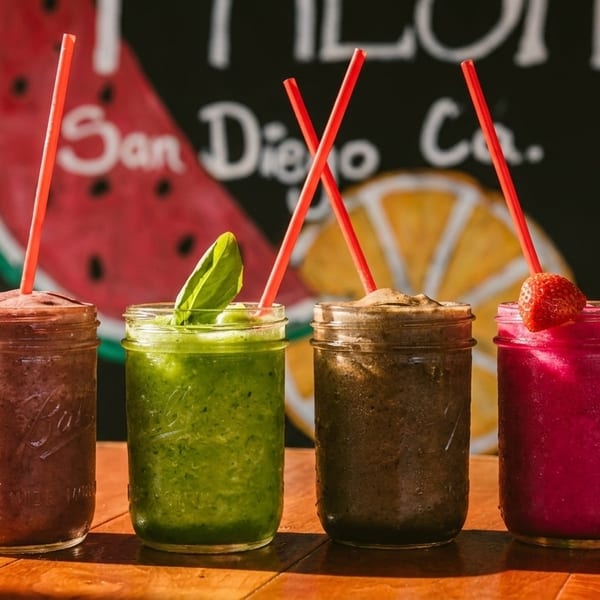 Four colorful smoothies in mason jars with red straws on a wooden table, against a fruit-themed backdrop.
