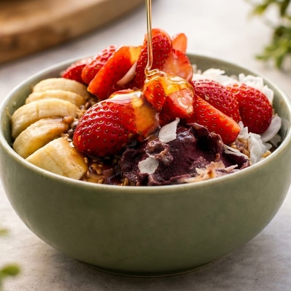 A green bowl of acai, bananas, strawberries, coconut flakes, and granola, with honey drizzled on top.