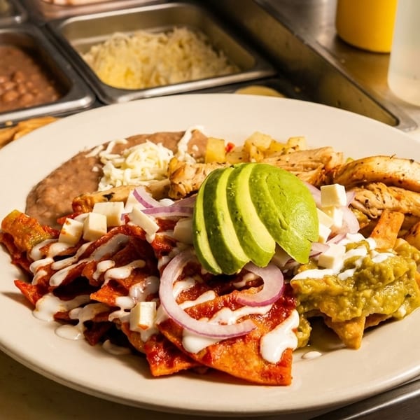 A plate of chilaquiles with red and green sauce, avocado, chicken, and refried beans.