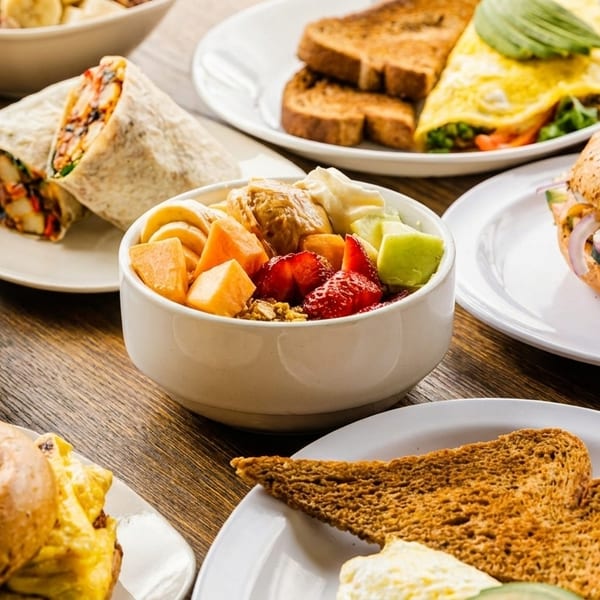 Table laden with breakfast items: omelet, toast, fruit bowl, bagel sandwiches, and a breakfast burrito.