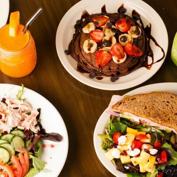 Overhead view of a table laden with two vibrant salads, a sandwich, chocolate pancakes, and two fresh juices.